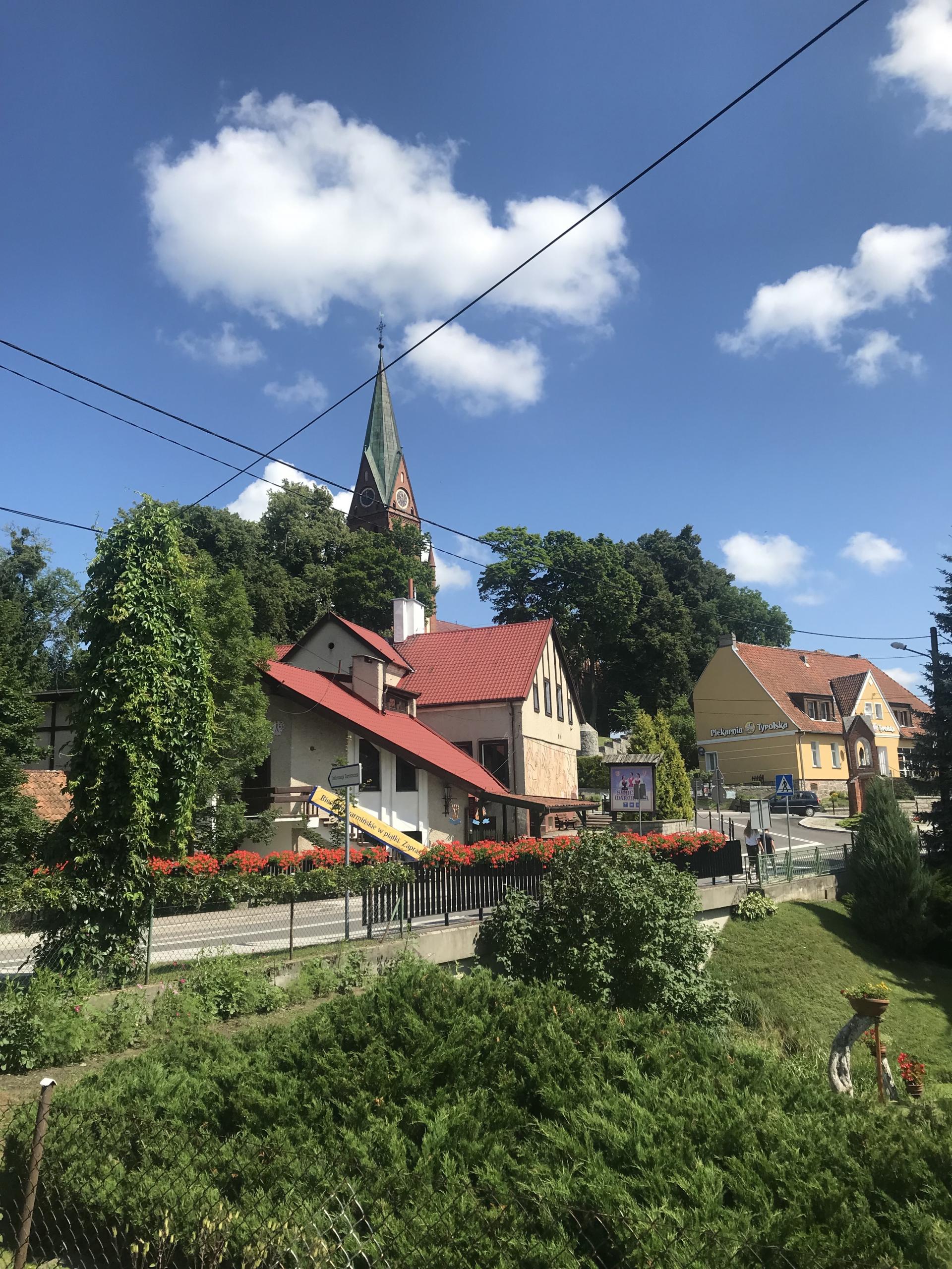 ulica w Gietrzwałdzie, przed nią duży pas zieleni, w tle zza drzew wyłania się wieża Sanktuarium Maryjnego