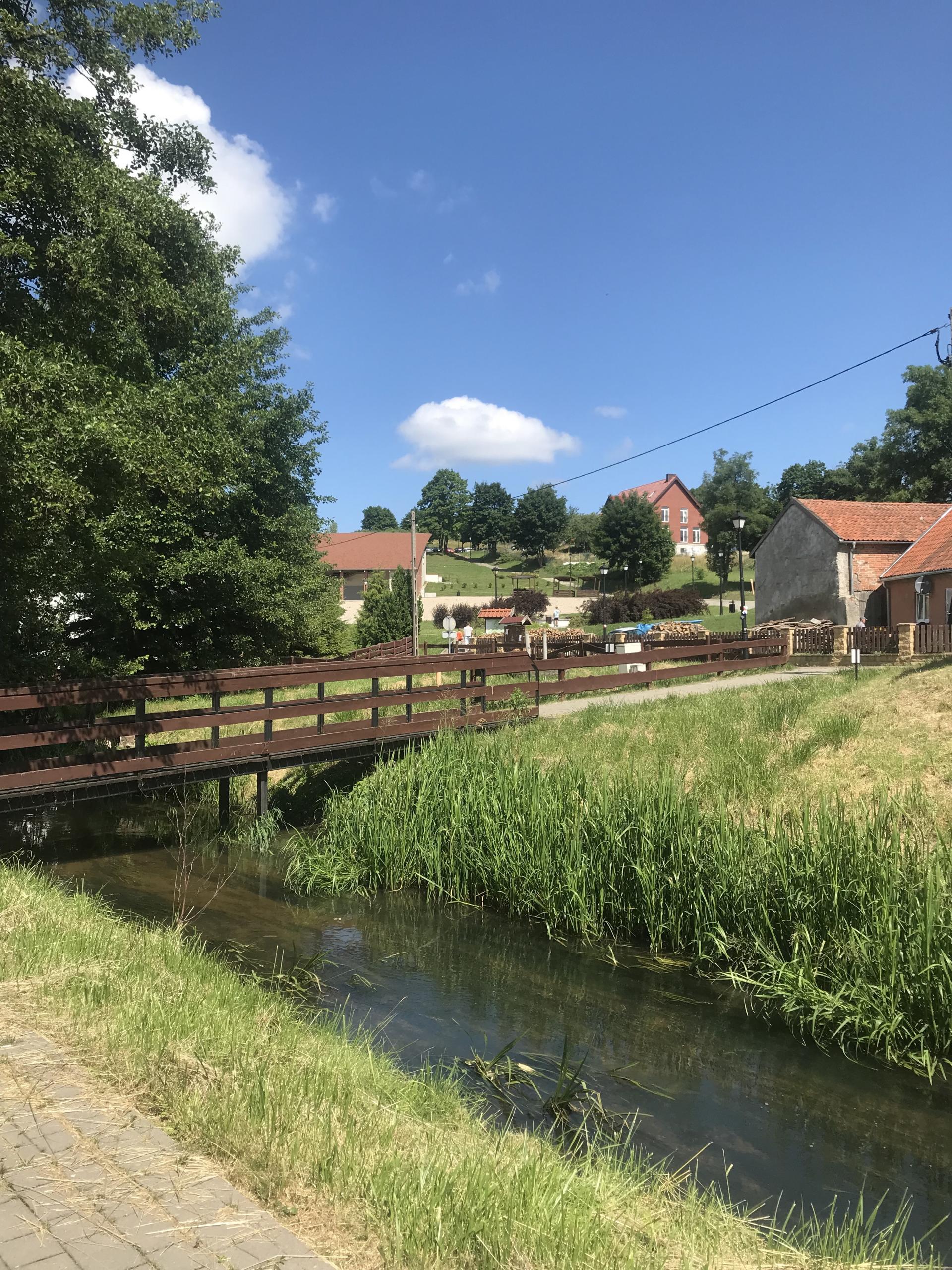 rzeczka, nad nią drewniany brązowy mostek, w tele zabudowania, widać trzy gospodarstwa